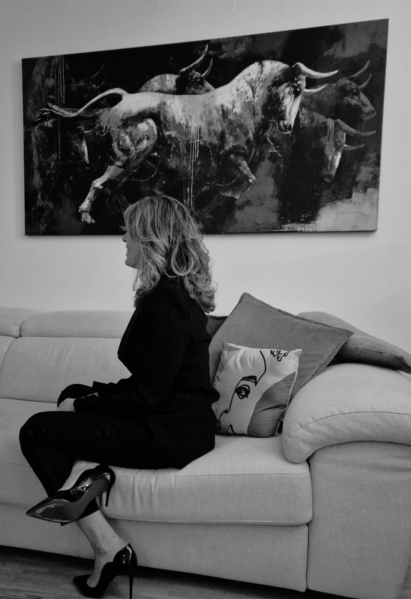 Femme assise sur un canapé sous un grand tableau mural, vêtue d'une robe noire et de talons hauts.