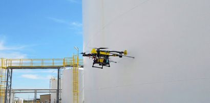 Drone spraying paint on a large white industrial tank beside a yellow scaffold under a blue sky