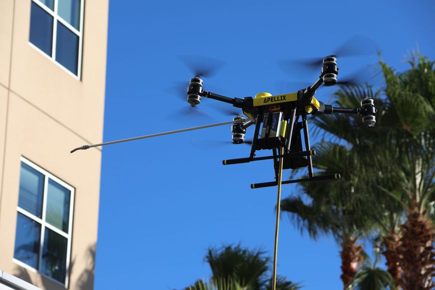 Drone flying beside a beige building and palm trees against a clear blue sky
