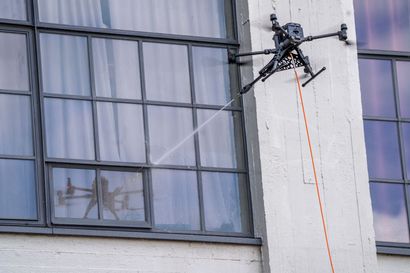 Drone hovering beside a building window, with a hanging orange tether line