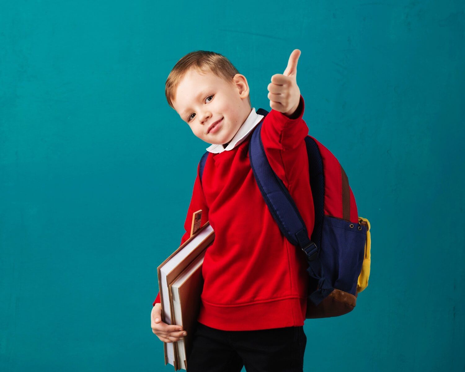 A young boy with a backpack is holding books and giving a thumbs up.