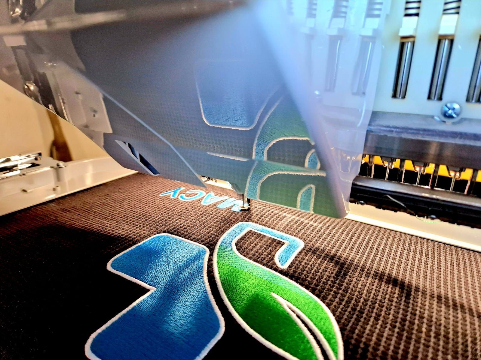 Embroidery machine stitching a logo onto dark fabric; blue, green, and white threads.