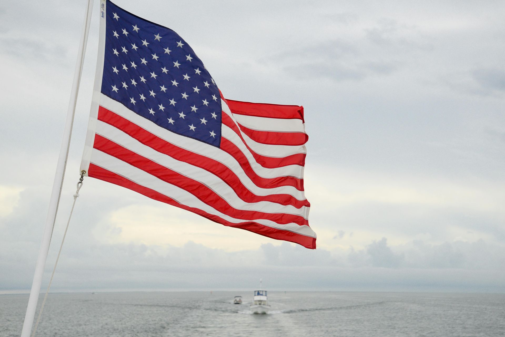 American flag waving on a pole, ocean and cloudy sky in the background, boat in the distance.