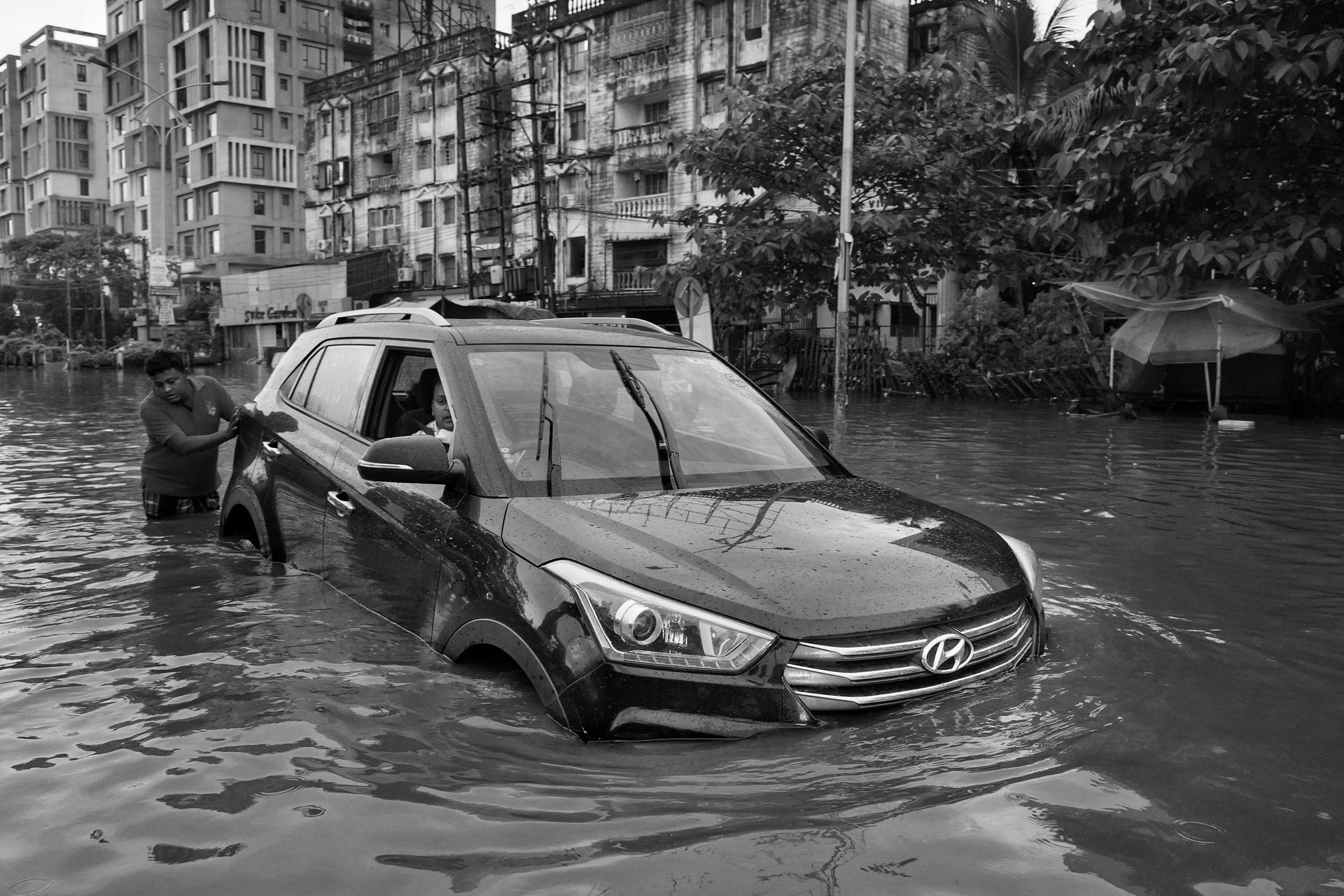 Car stranded in flooded street; man pushing from the side in front of buildings.