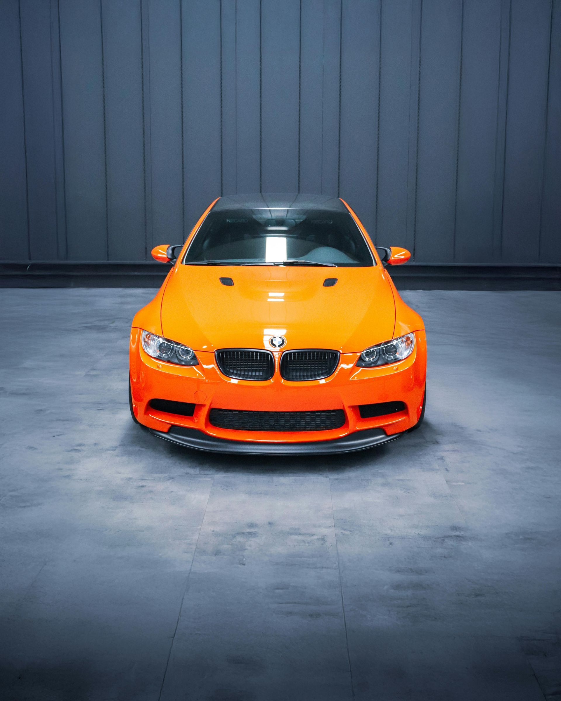 Bright orange BMW M3 sports car, centered against a dark gray wall.