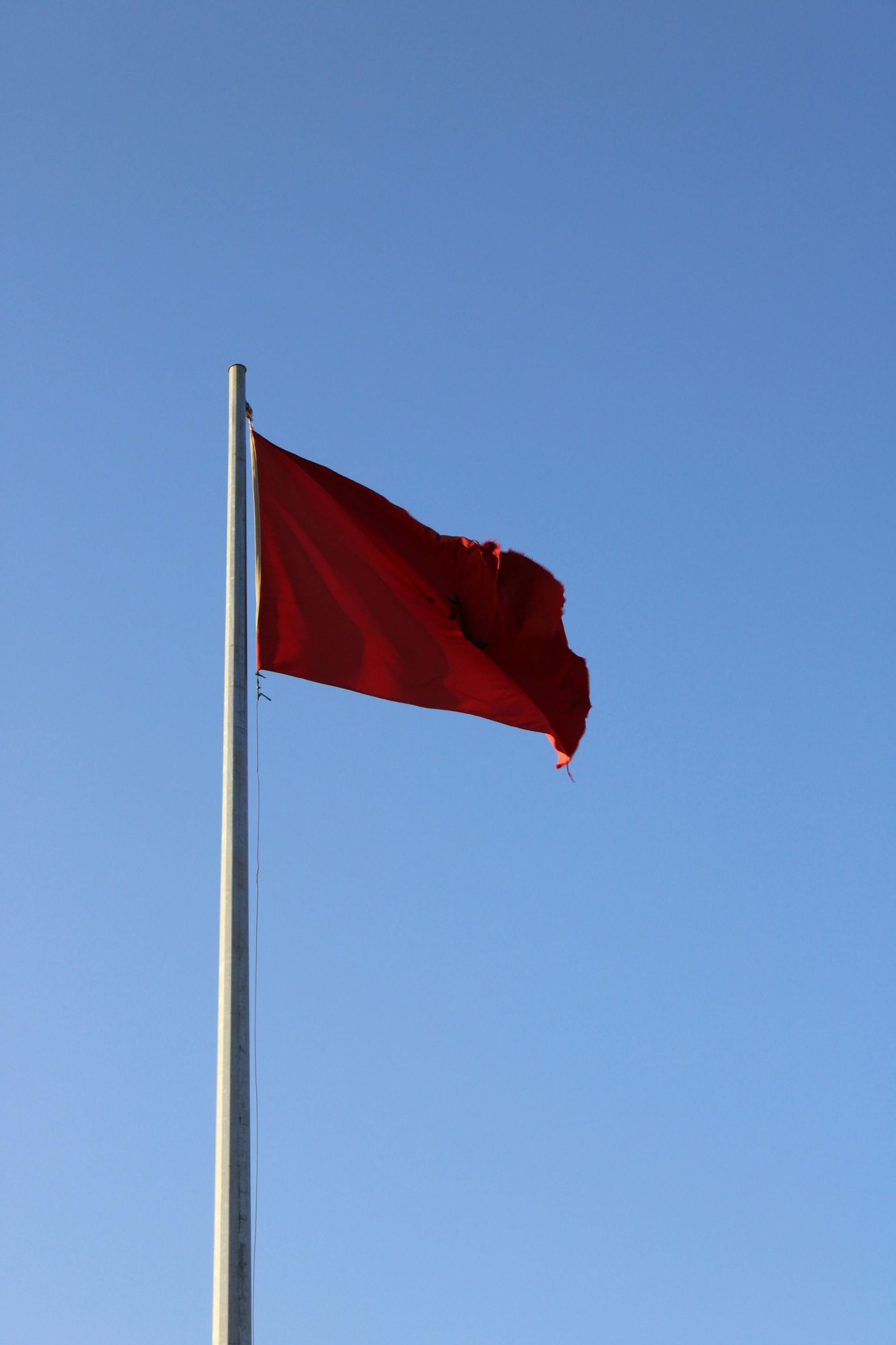 Red flag waving on a silver pole against a bright blue sky.
