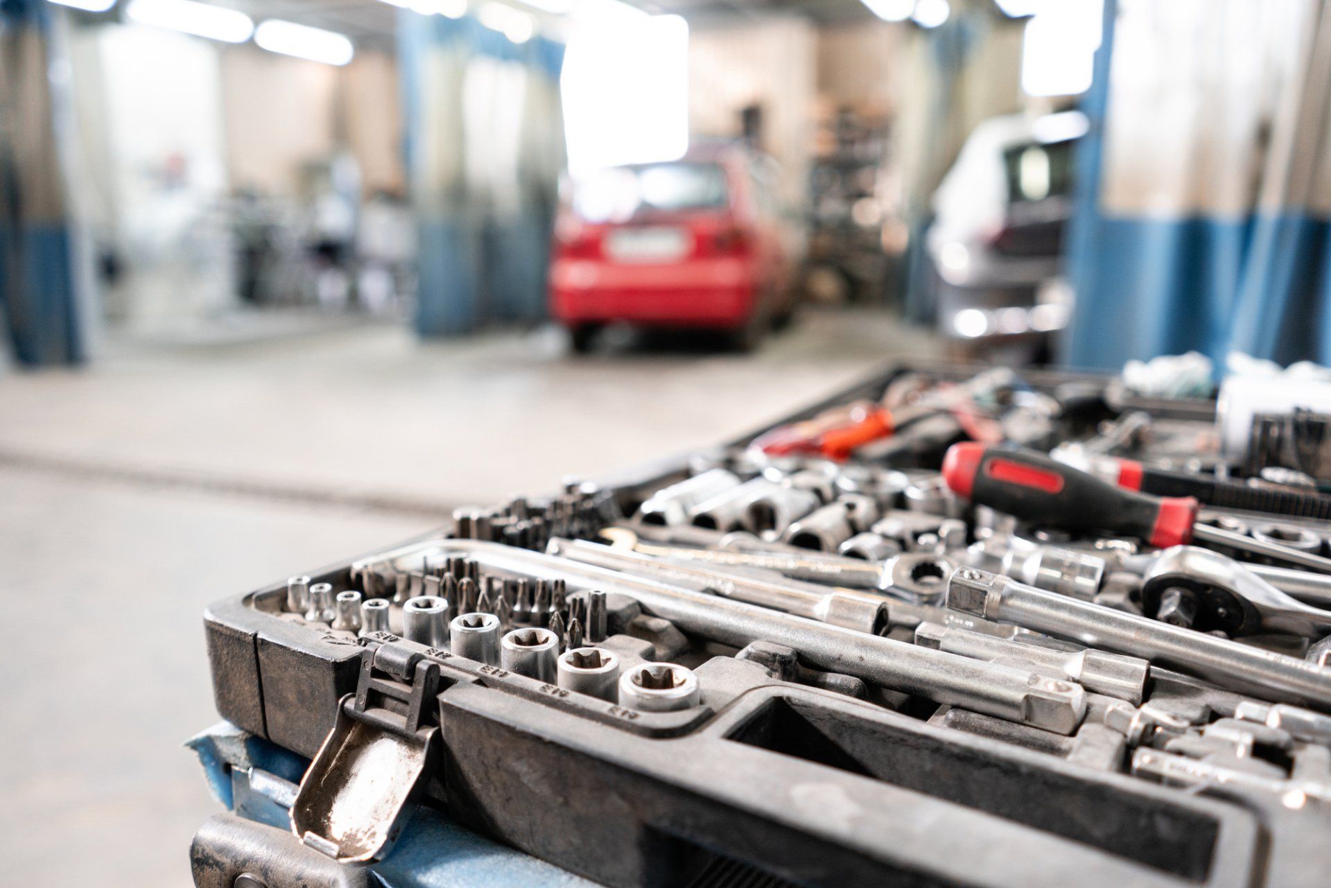 A close-up of a mechanic's tool kit in the foreground, with a blurred auto repair shop and red car in the background.