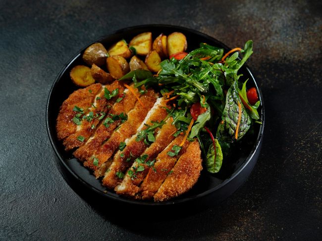 Eine schwarze Schüssel gefüllt mit gebratenem Hühnchen, Kartoffeln und Salat.