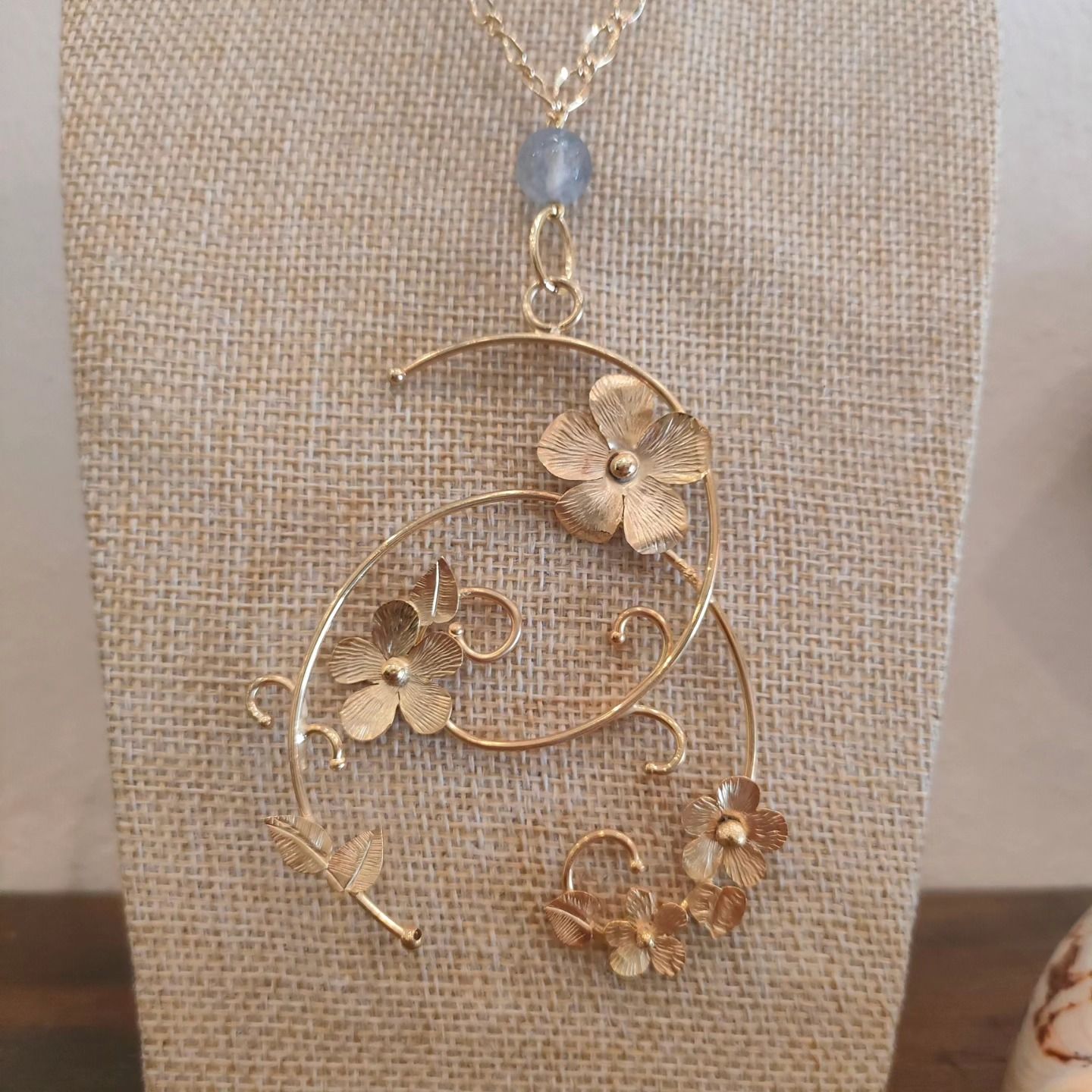 Collana con ciondolo in filo metallico dorato a forma di fiore, con fiorellini e una perlina blu, esposta su un busto in lino