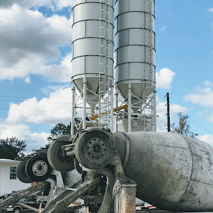 Two tall, gray industrial silos stand behind a concrete mixer truck against a blue sky with white clouds.