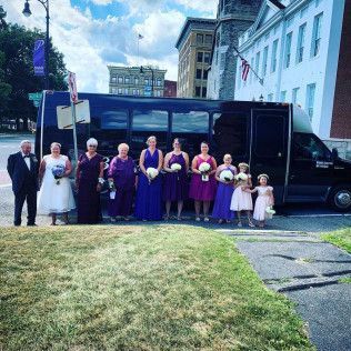 A wedding party stands outside a large black passenger bus parked on a city street near historic brick buildings.