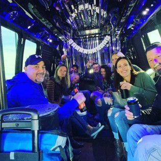 A group of people laughing and holding drinks while sitting inside a festive party bus with blue lighting and decor.