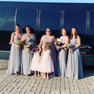 Seven people pose in formal wear in front of a dark bus, including two children in white dresses and five in gray gowns.