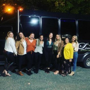 A group of eight people stand in a line outside a dark limo bus at night, smiling together.