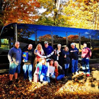 A group of 11 people stand smiling together in front of a parked bus surrounded by autumn leaves.