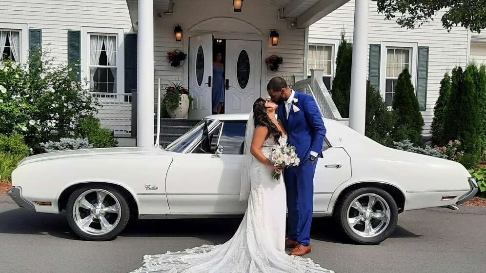 A bride in a white wedding gown and a groom in a blue suit kiss in front of a white classic car at a building entrance.