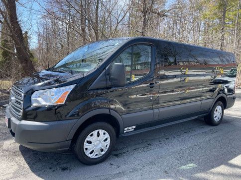 A shiny, black Ford Transit cargo van parked on an asphalt lot with bare trees in the background.