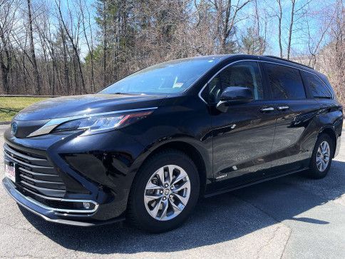 A shiny black Toyota Sienna minivan parked on an asphalt lot with a wooded background.