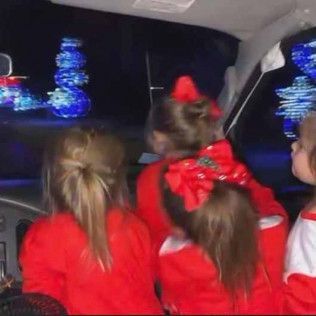 Three children in red shirts sit in a car looking out at glowing blue holiday light displays in the dark.