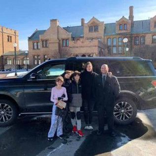 Four people pose in front of a large black SUV parked in front of a historic, multi-story stone building.