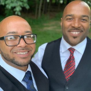 Two men smile side-by-side, wearing dress shirts, ties, and vests, with an outdoor greenery background.