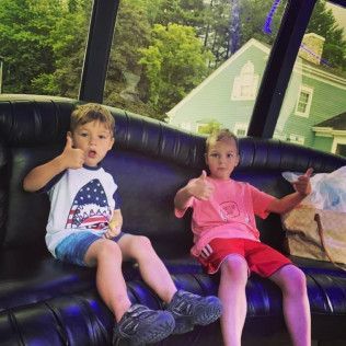 Two children sit on a black leather limousine bench, smiling and giving a thumbs-up gesture.