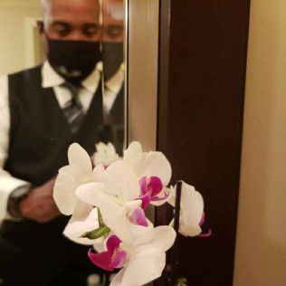 A person wearing a mask, white shirt, and vest reflects in a mirror behind a cluster of white orchids with purple centers.