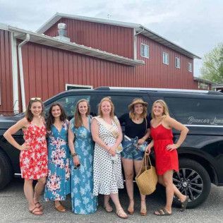 Six friends stand smiling in front of a black SUV parked beside a red metal building.