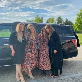 Four people stand in a line smiling in front of a parked black vehicle on a paved surface during the day.