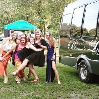 Five people pose playfully together on grass in front of a dark bus, with a green canopy tent visible in the background.