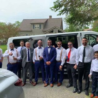 A group of people in semi-formal wedding attire stand outdoors in front of a white passenger van.