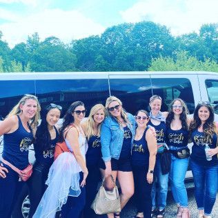 Ten people wearing matching navy tank tops stand in a line smiling in front of a white passenger van on a sunny day.