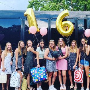 A group of teenagers stand in front of a dark bus with large gold “16” balloons, holding gifts and party decorations.