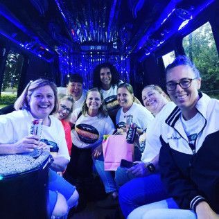 Seven people smiling inside a dimly lit vehicle with blue neon lights, some holding beverage cans and a gift bag.