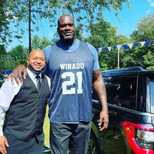 Shaquille O’Neal stands next to a smiling man in a suit vest outdoors with a car in the background.
