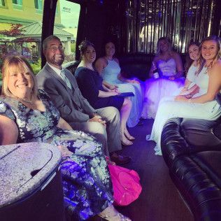A group of people dressed in formal attire sit inside a limousine, with some holding drinks and smiling at the camera.
