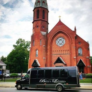 A black shuttle bus parked in front of a tall, red brick church with a steeple and a rose window under a cloudy sky.