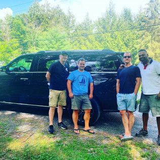 Four people stand in a line posing in front of a black SUV parked on a gravel path near a forest.
