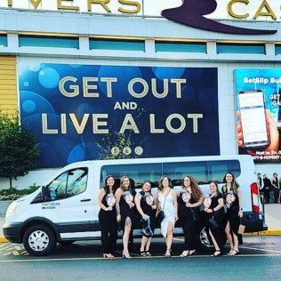 A group of eight people posing in front of a white passenger van parked outside a casino with a large promotional sign.