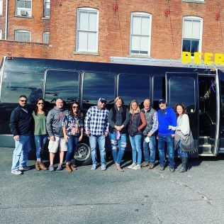A group of twelve people stand in a line posing in front of a parked black bus outside a red brick building.