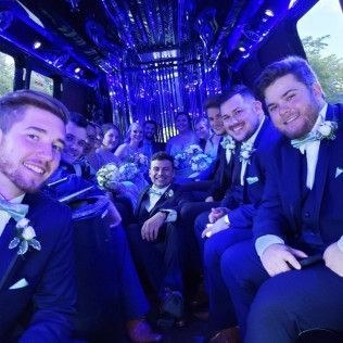 A group of people in formal attire, some wearing boutonnieres, sit inside a brightly lit party bus with blue lighting.