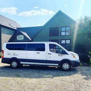 A white passenger van with a branded logo parked in front of a green building on a gravel lot under a blue sky.