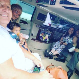 A family rides in a limousine. A person in the foreground takes a selfie while a child and two others sit in the back.