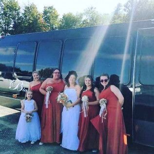 A wedding party poses for a photo in front of a black limousine, with one person in white and five in dark orange dresses.