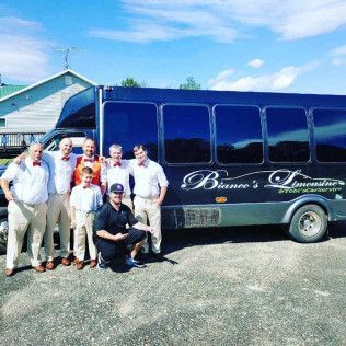 A group of people in formal attire and one man in a black shirt pose in front of a dark blue Bianco's Limousine bus.