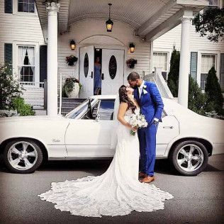 A bride in a white wedding gown and a groom in a blue suit kiss in front of a white vintage car parked at a building.