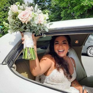 A smiling bride in a white wedding dress sits in a car, holding a bouquet of light pink roses and greenery out the window.