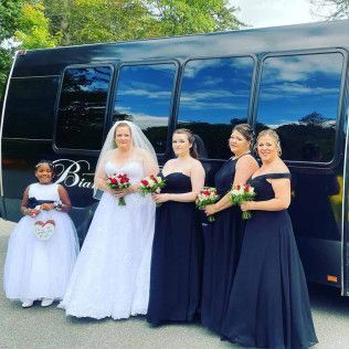 A bride, three bridesmaids in navy gowns, and a child in a white dress pose in front of a black limousine.