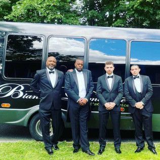 Four men in black tuxedos stand in front of a parked black bus on a grassy area.