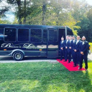 Six men in suits stand in a line on a red carpet leading into a black party bus parked on grass.